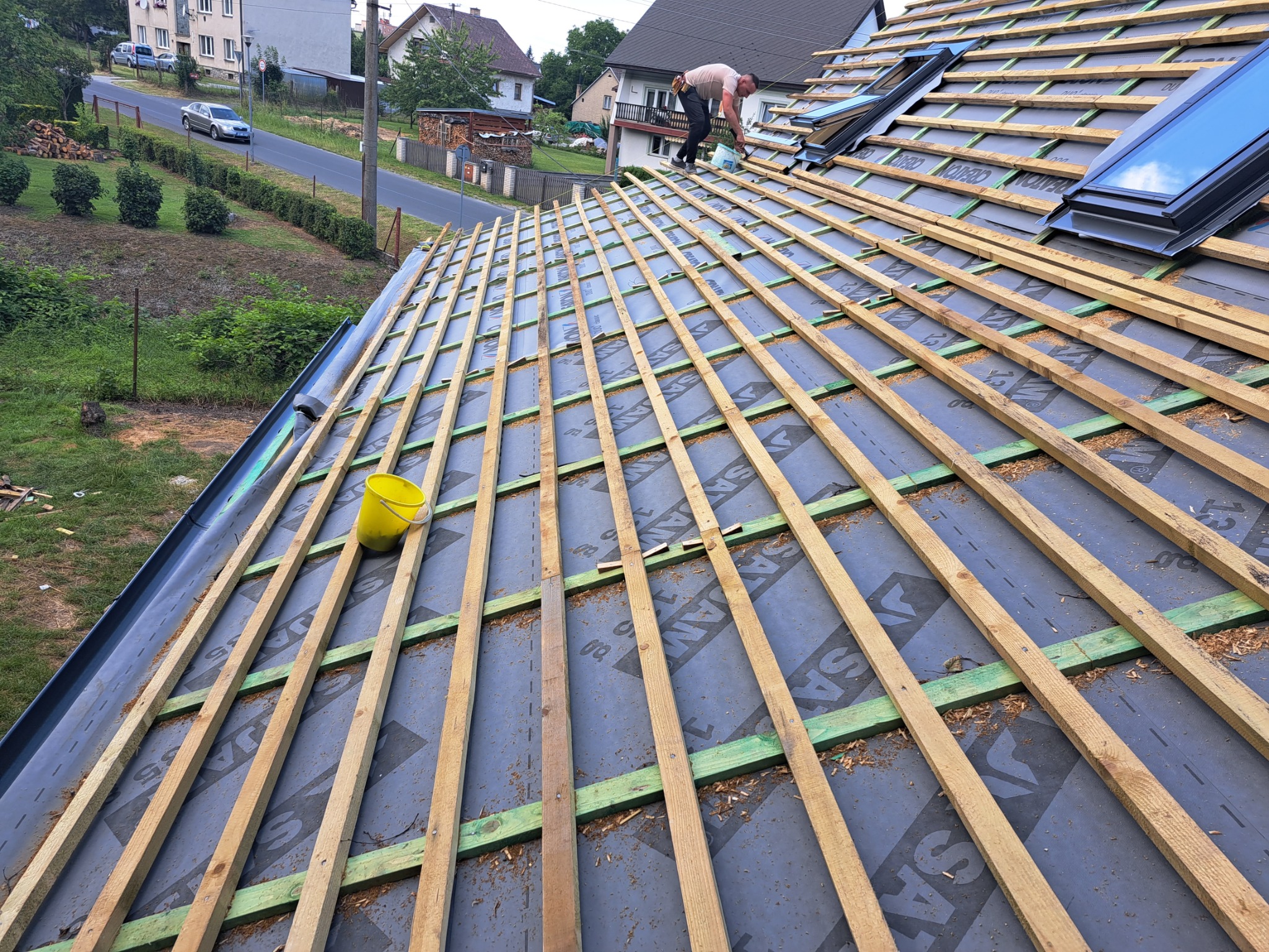 Montaż łat dachowych na membranie z widocznymi oknami dachowymi i żółtym wiadrem, w tle domy jednorodzinne i ulica.