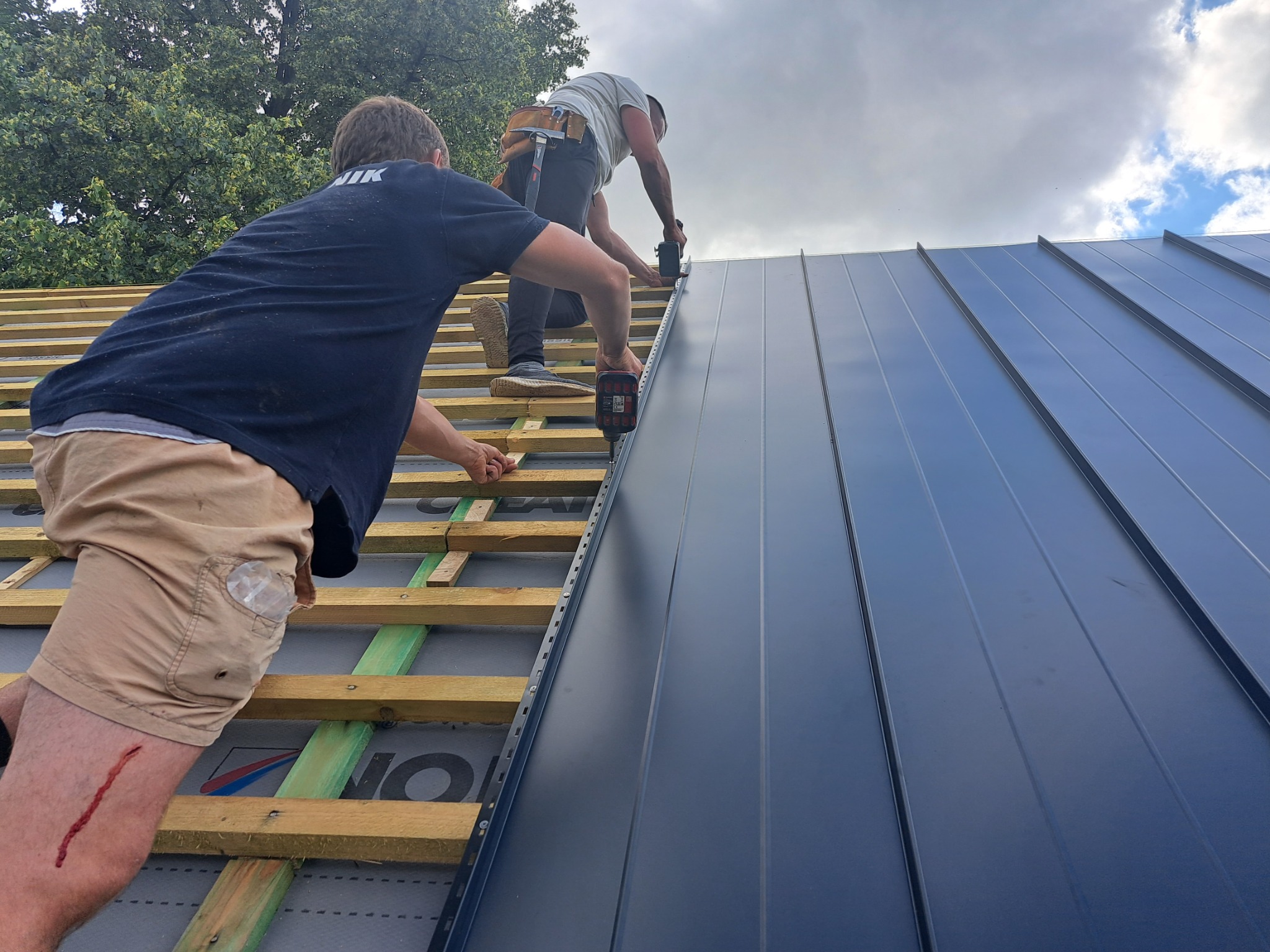 Dwóch dekarzy montuje ciemnoszare panele na drewnianej konstrukcji dachu; jeden z nich ma ranę na nodze.