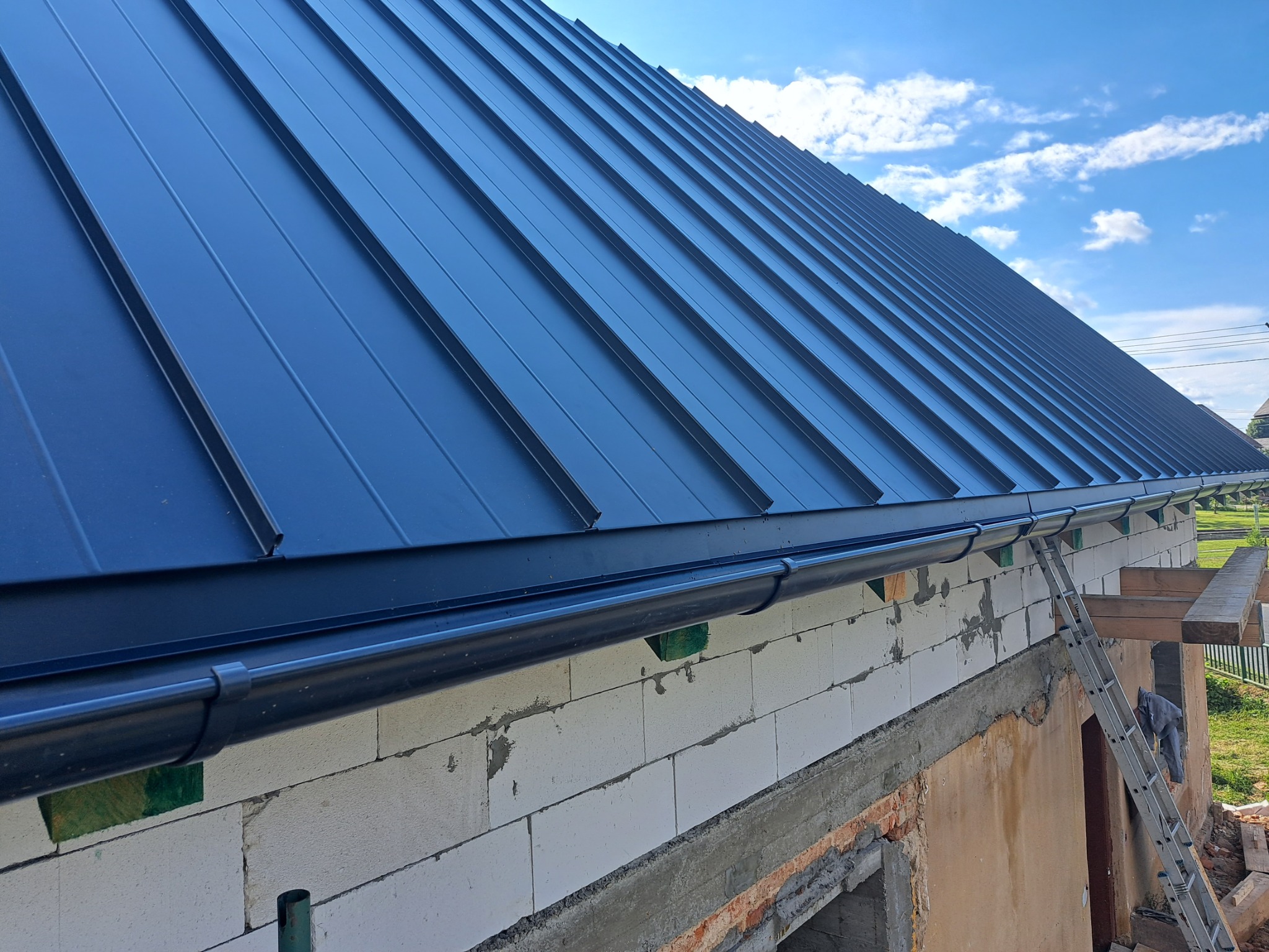 Nowe, czarne, metalowe pokrycie dachowe z widocznymi rynnami, zamontowane na budynku w trakcie budowy, z widocznymi bloczkami betonowymi i rusztowaniem.