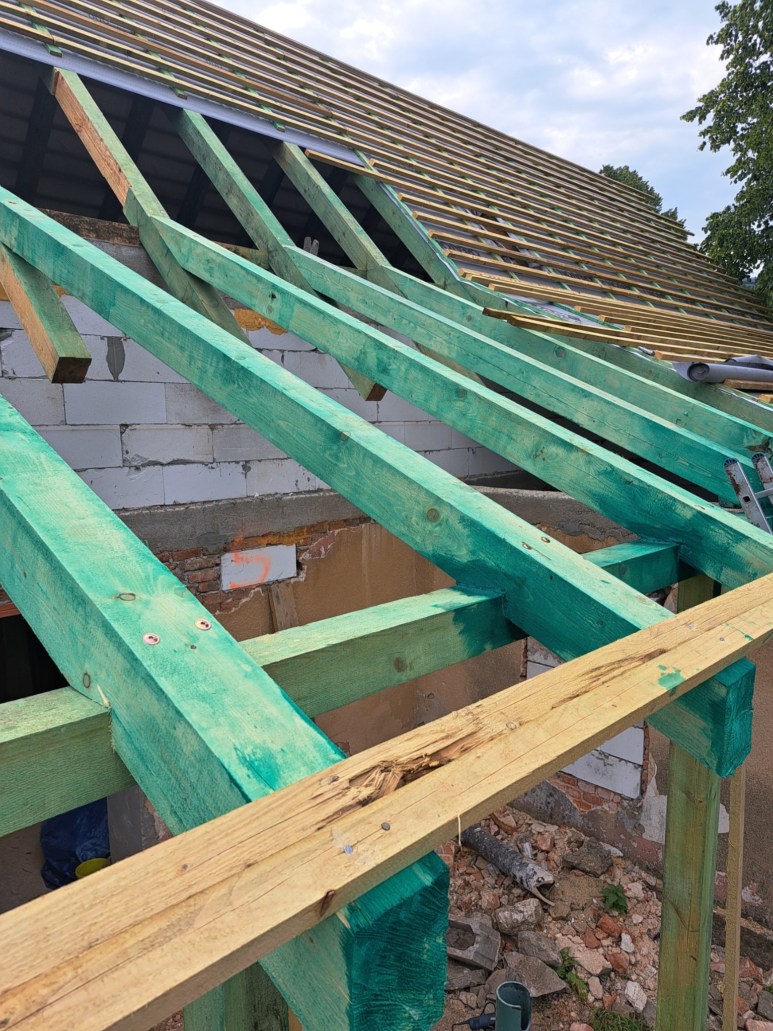 Konstrukcja drewniana dachu z zielonymi belkami, widok na łatowanie i niebo, fragment ściany z cegły w tle.