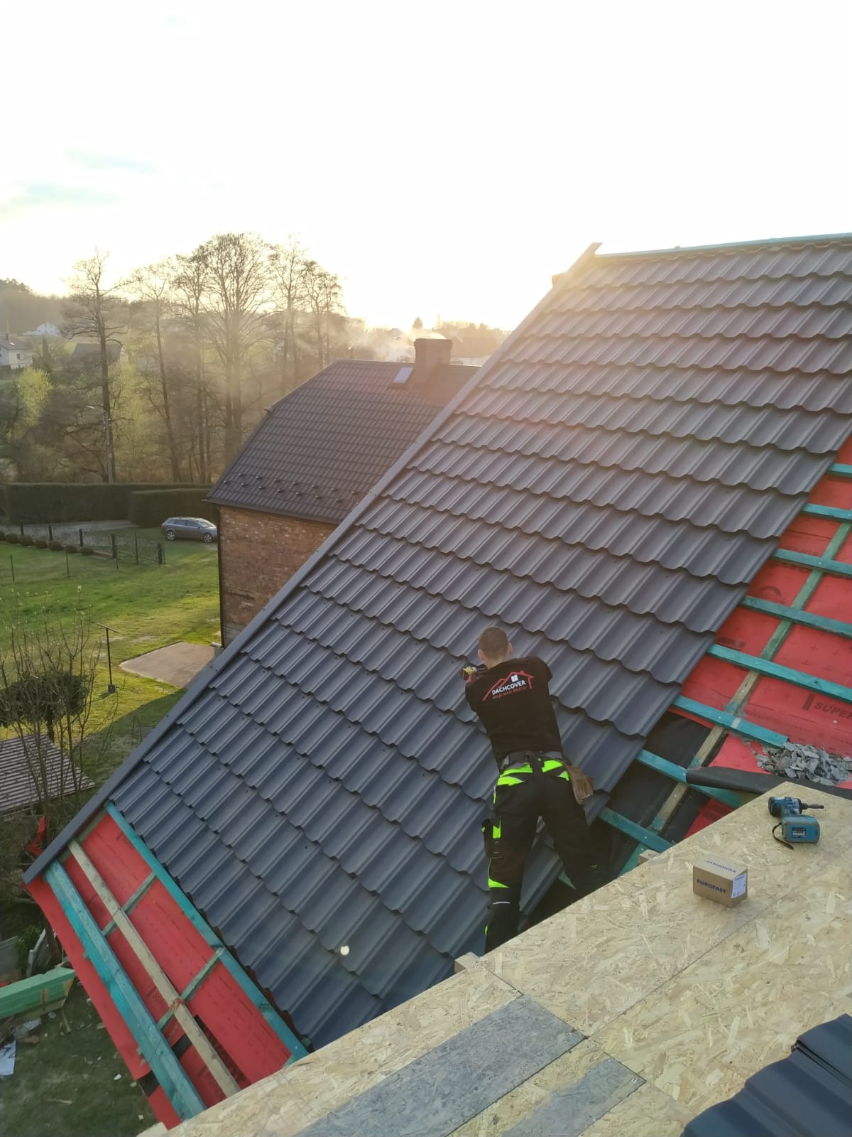 Dekarz montuje grafitową dachówkę na nowym dachu, widoczne narzędzia i elementy konstrukcji dachu na tle zabudowań i zieleni.