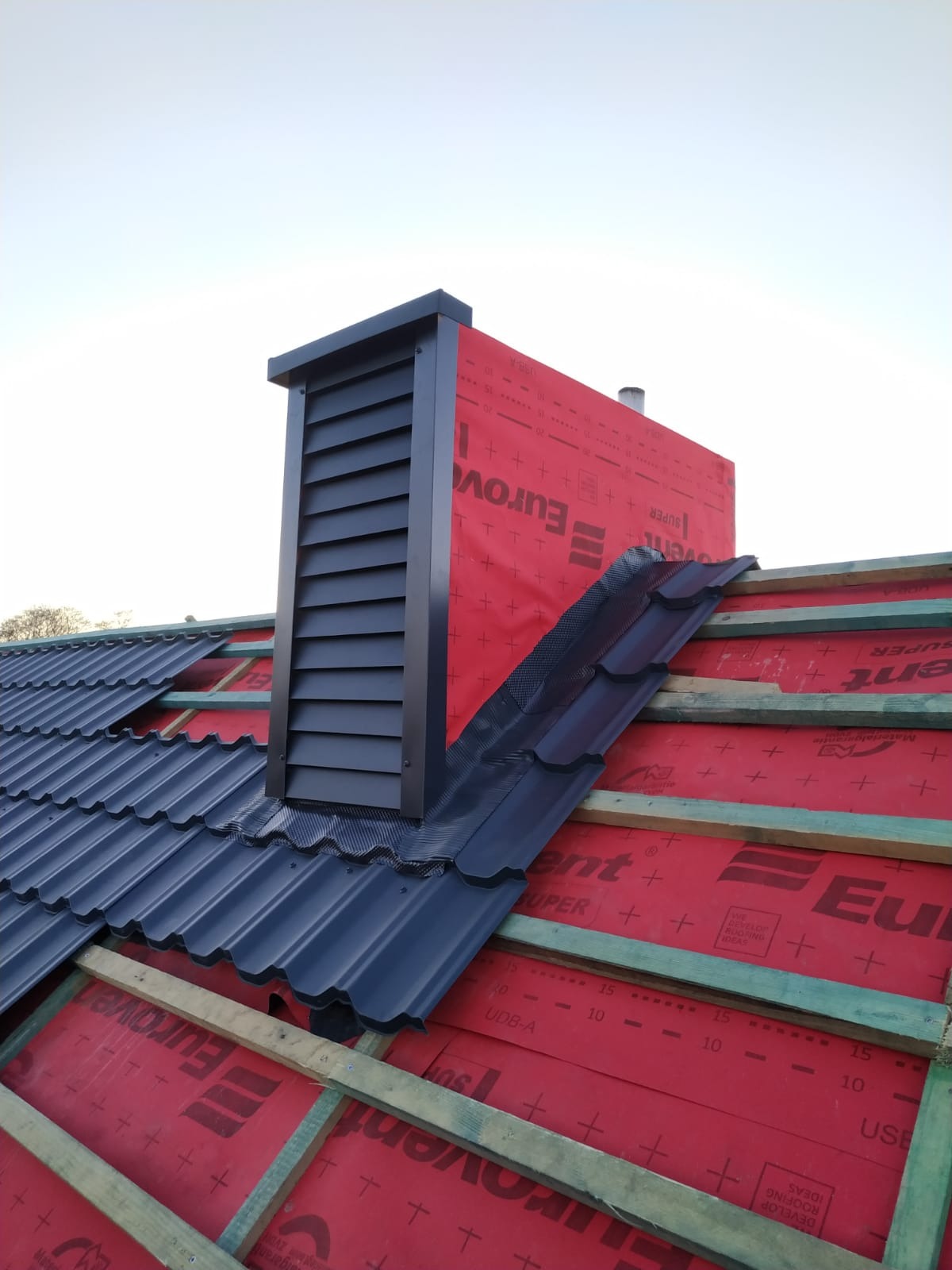 Fragment dachu w trakcie krycia z widocznym kominem wentylacyjnym, deskami i membraną dachową w kolorze czerwonym.