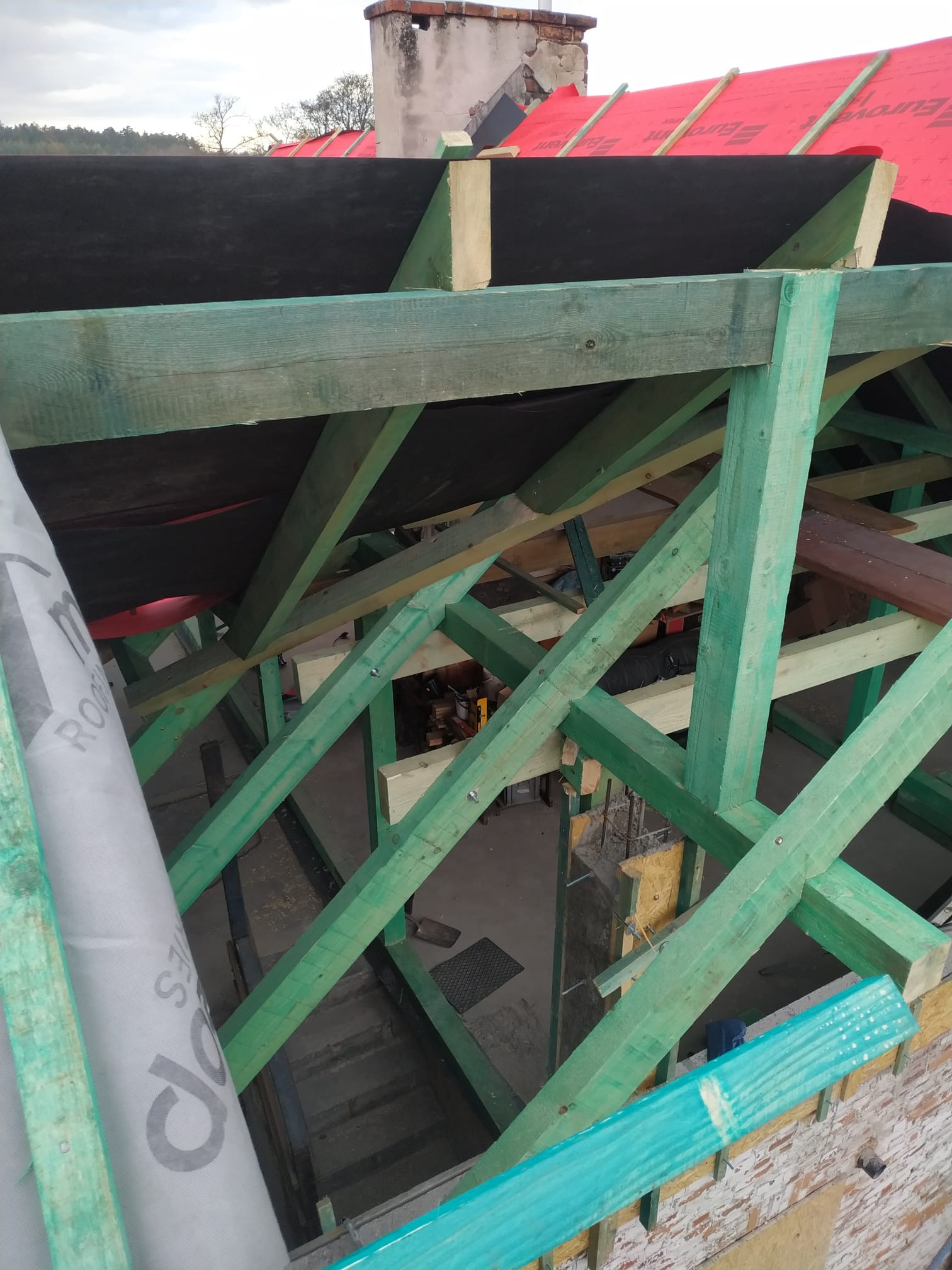 Konstrukcja dachu z impregnowanego drewna, widoczna czarna membrana dachowa i fragment czerwonej folii dachowej, w tle komin i pochmurne niebo.