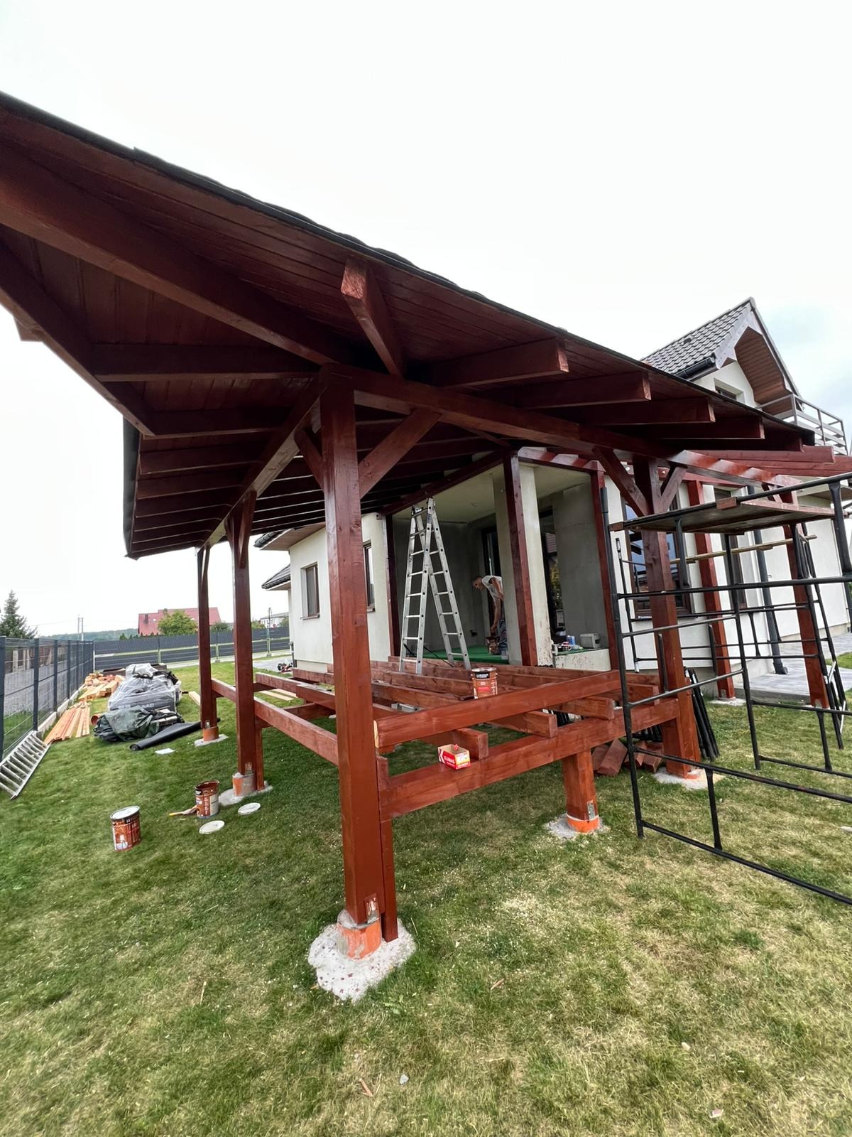 Drewniana konstrukcja z dachem w trakcie budowy, pomalowana na brązowo, z widocznymi puszkami farby i drabiną. W tle dom jednorodzinny i zielony trawnik.