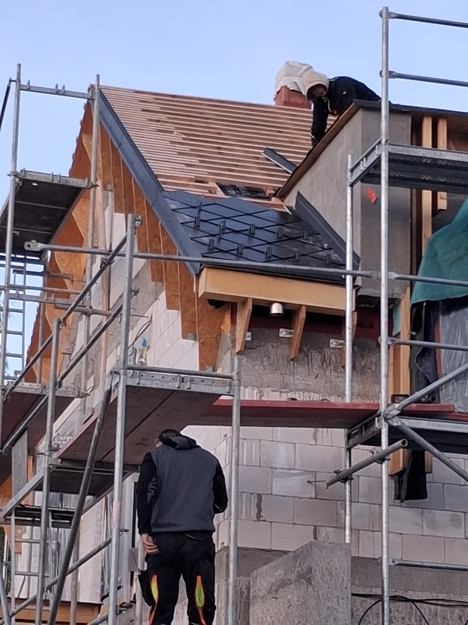 Montaż dachu z czarnej dachówki karpiówki na drewnianej konstrukcji, widoczne rusztowania i pracownicy. Częściowo pokryty dach z drewnianą łatą. Ujęcie z dołu.