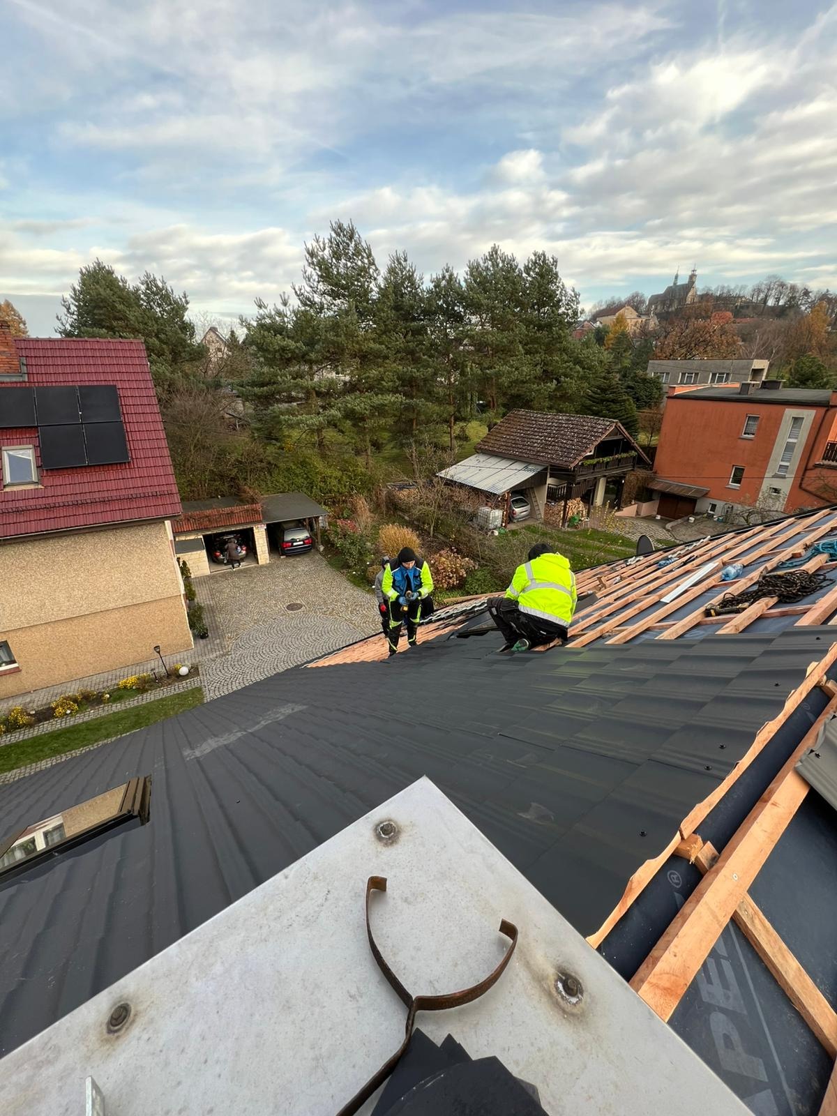Montaż czarnej blachodachówki na dachu domu jednorodzinnego. Dwóch dekarzy w odblaskowych ubraniach pracuje na wysokości. Widok z góry na ukończony fragment dachu.