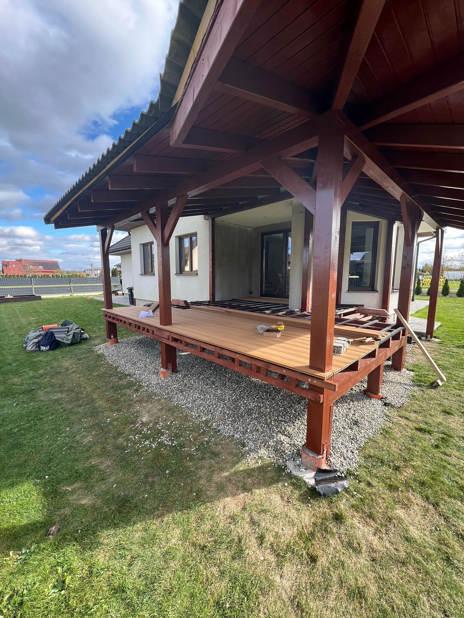 Wznoszenie drewnianej konstrukcji dachu z brązowymi słupami i szarym pokryciem na tle domu jednorodzinnego i zielonego trawnika w słoneczny dzień.