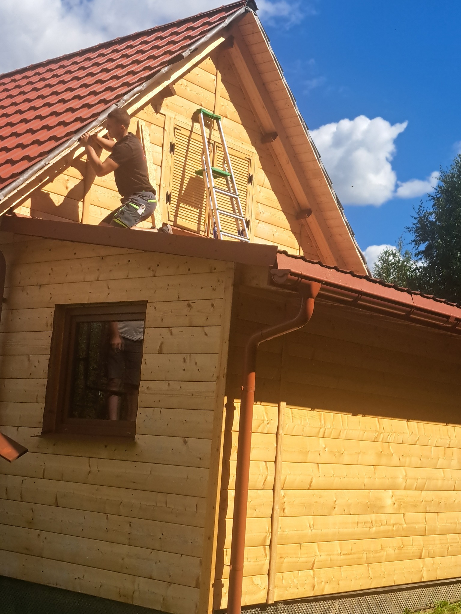 Praca na dachu drewnianego domu z czerwoną dachówką. Mężczyzna montuje elementy, obok drabina. Widoczne rynny i błękitne niebo z chmurami.