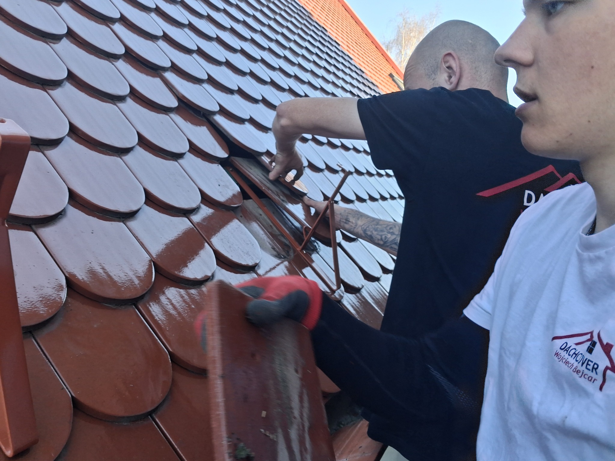 Ekipa montuje brązową dachówkę karpiówkę na dachu. Widoczne ręce w rękawicach, fragment koszulki z logo firmy dekarskiej i fragment dachu sąsiada.