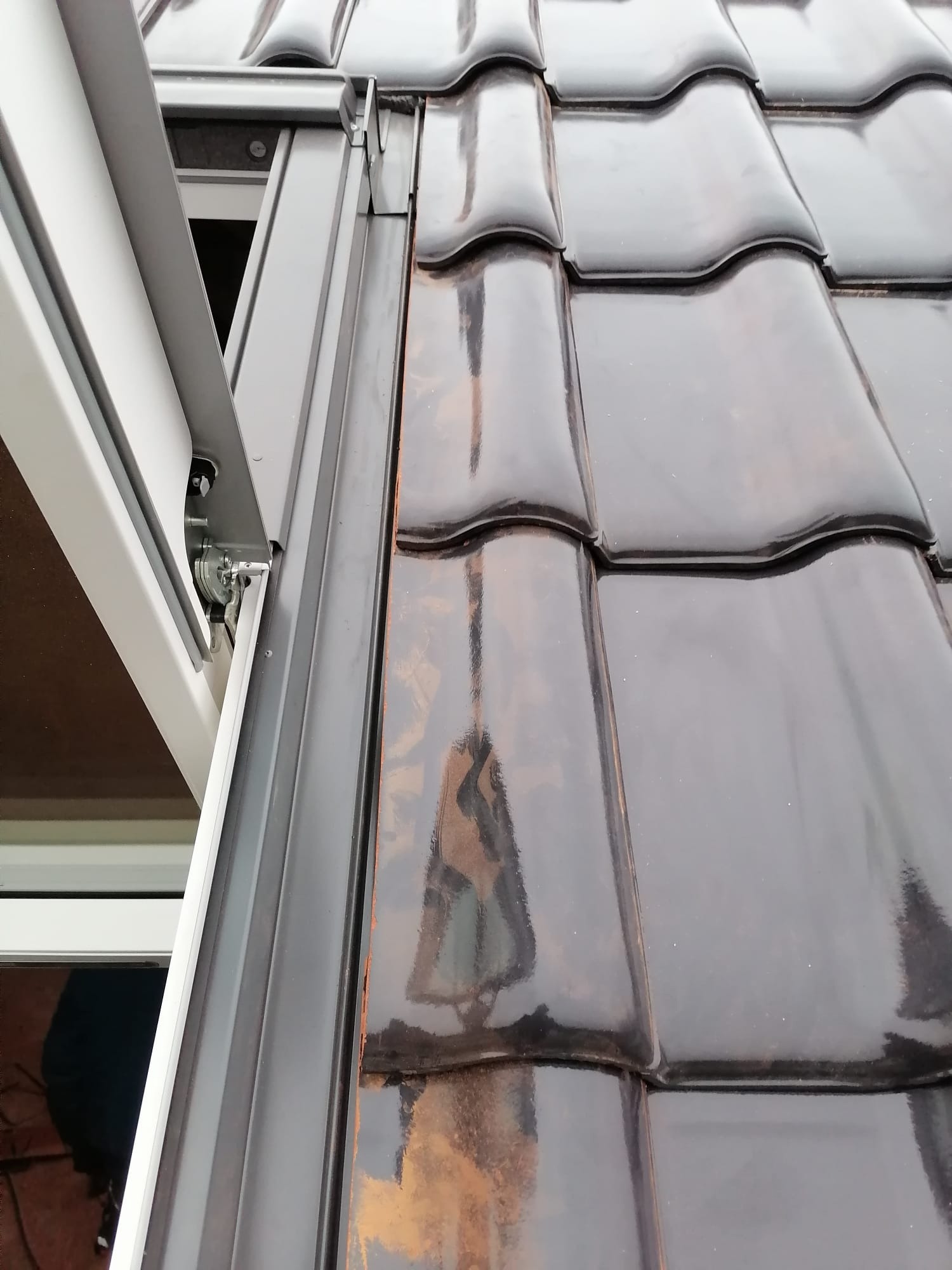 Błyszcząca, brązowa dachówka ceramiczna z oknem dachowym i obróbką blacharską w kolorze grafitowym, widok z bliska.