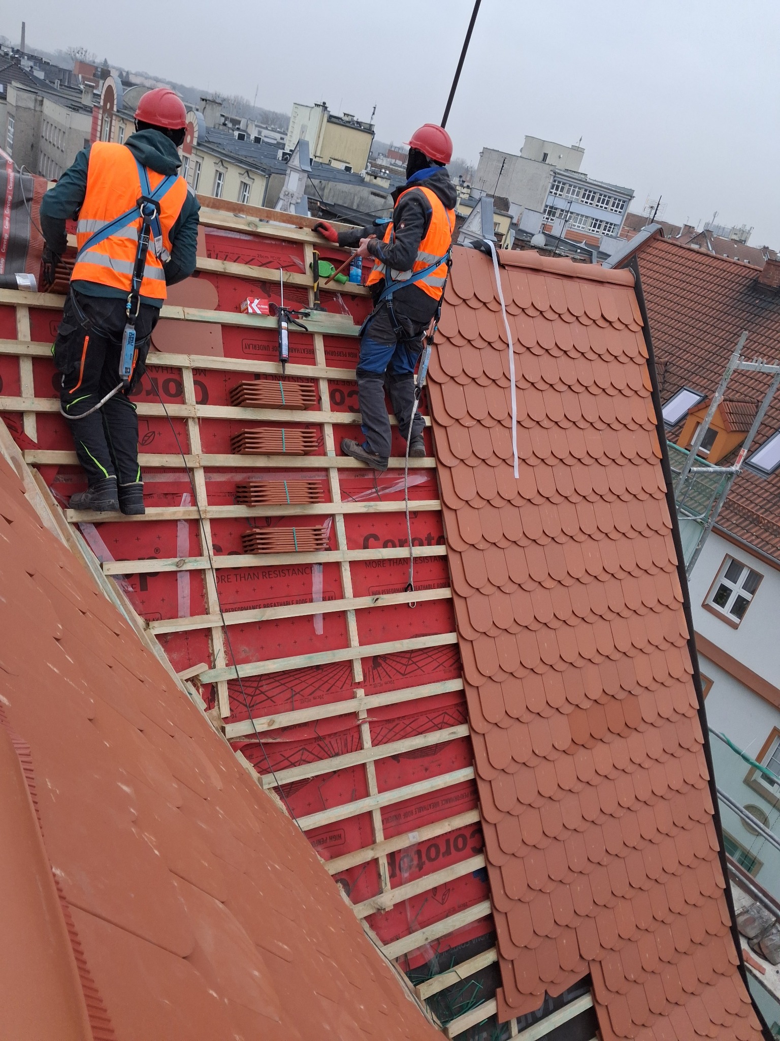 Dekarze w pomarańczowych kamizelkach montują czerwoną dachówkę na drewnianej konstrukcji dachu. Widok z góry na częściowo pokryty dach i zabudowę miejską w tle.