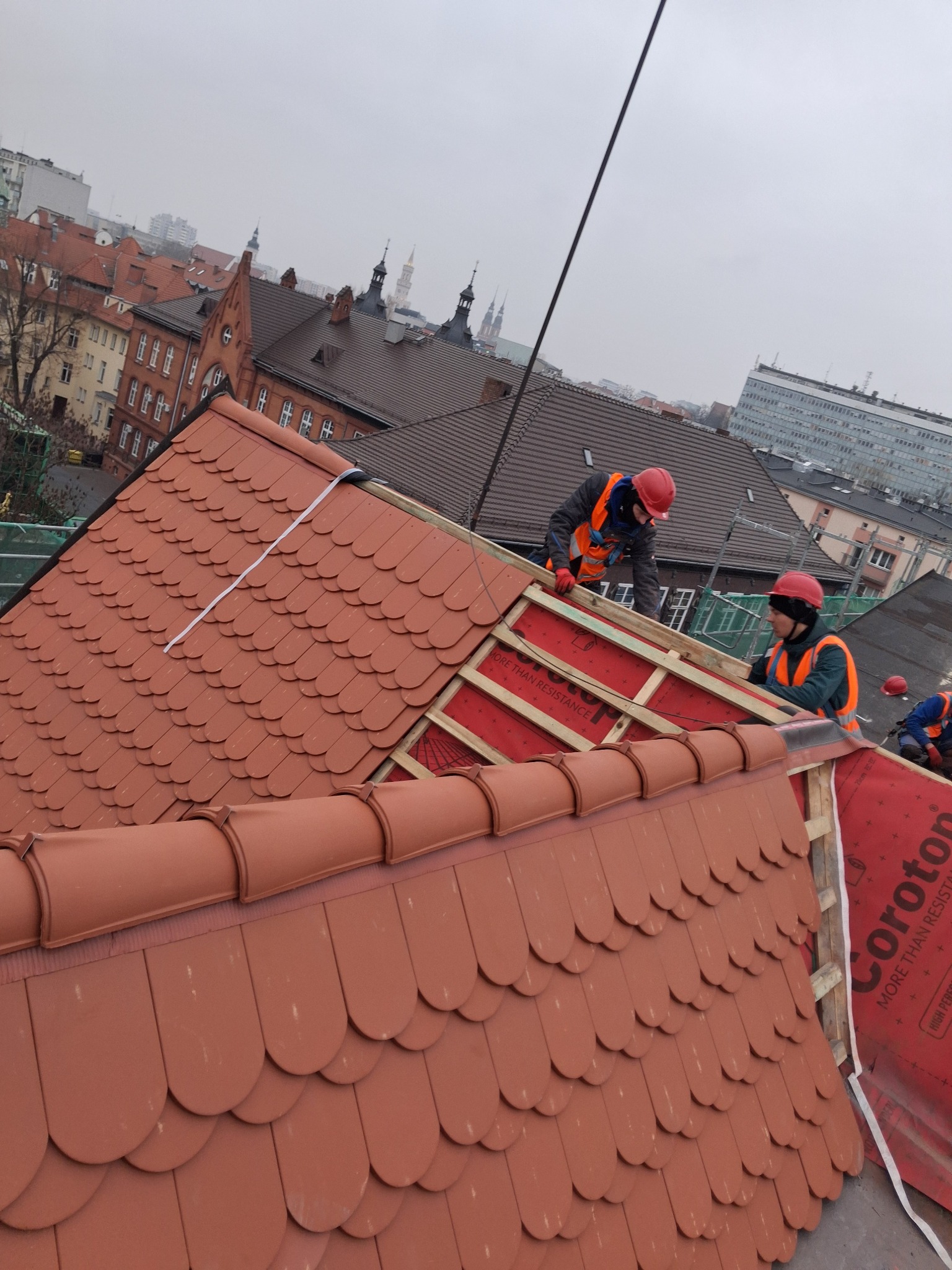 Montaż dachówki karpiówki na dachu budynku. Widoczni dekarze w kaskach i kamizelkach. W tle zabudowa miejska i pochmurne niebo. Prace dekarskie w toku.