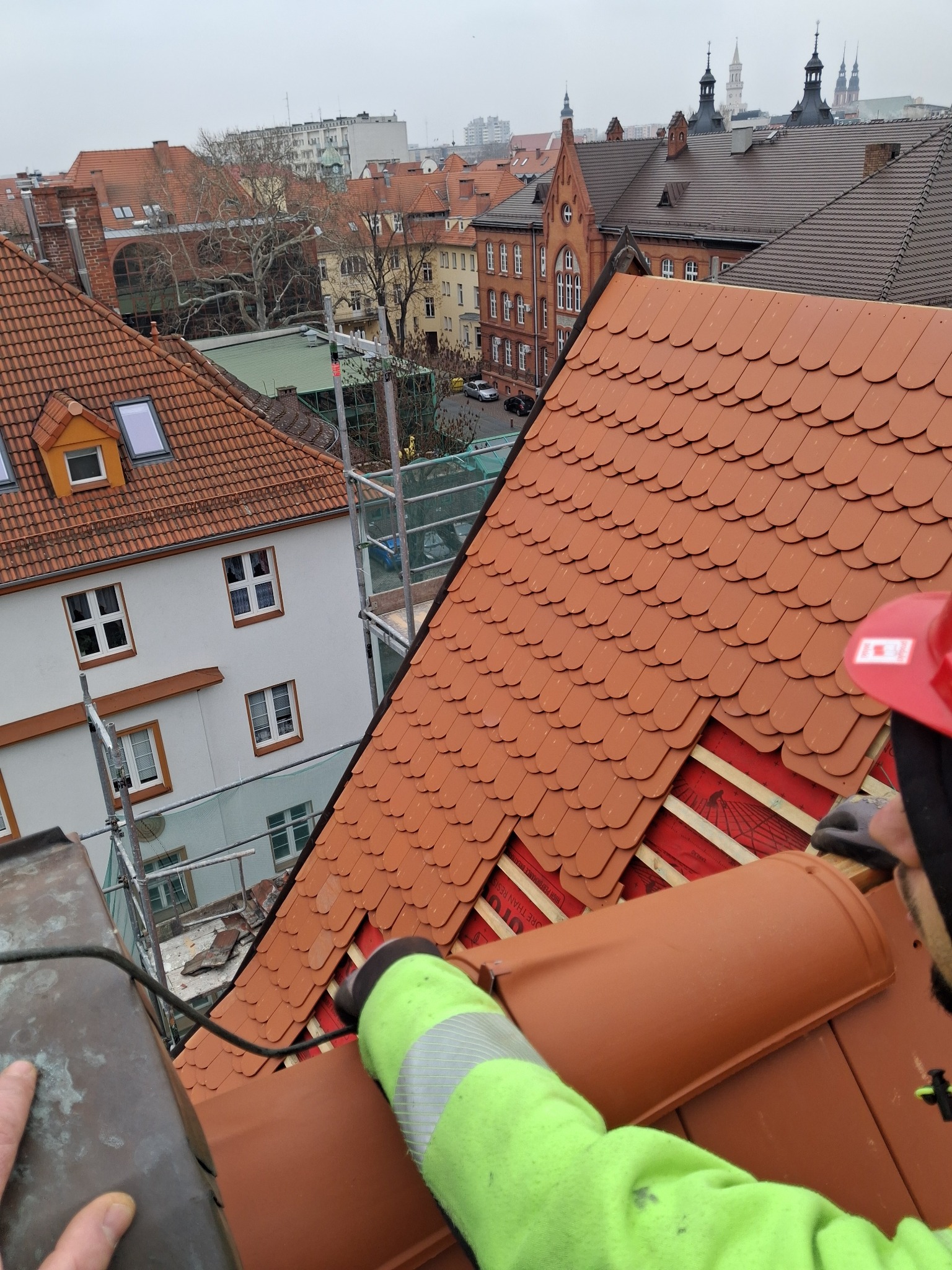 Układanie dachówki karpiówki na dachu budynku. Widoczne elementy konstrukcji dachu i fragment sylwetki dekarza w odblaskowej kurtce. W tle zabudowa miejska.