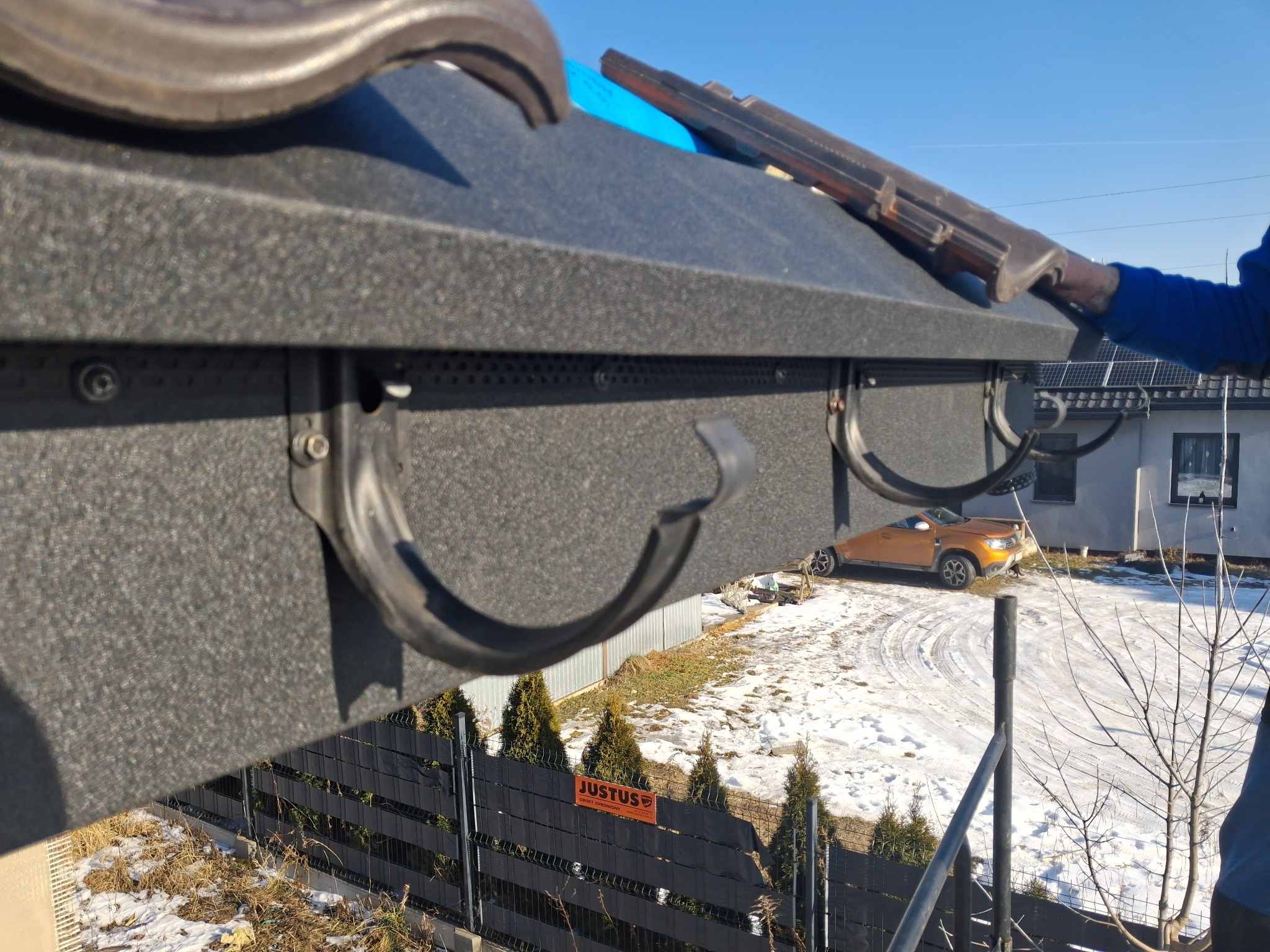 Montaż rynny na dachu z blachy trapezowej, widok z góry na częściowo ośnieżony teren i dom w tle. Widoczny fragment ręki dekarza w niebieskiej bluzie.