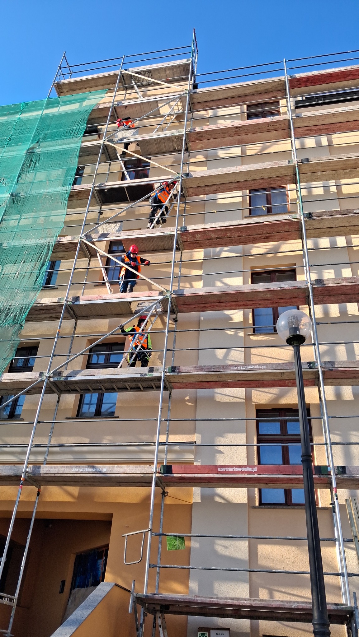 Pracownicy na rusztowaniu przy budynku z oknami, zabezpieczonym siatką. Widok z dołu, błękitne niebo w tle. Praca na wysokości, ochrona osobista.