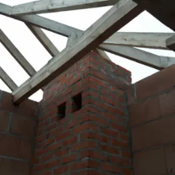 Wznoszenie komina z cegły klinkierowej na tle drewnianej konstrukcji dachu i ścian z pustak&oacute;w ceramicznych. Widoczne otwory wentylacyjne w kominie.