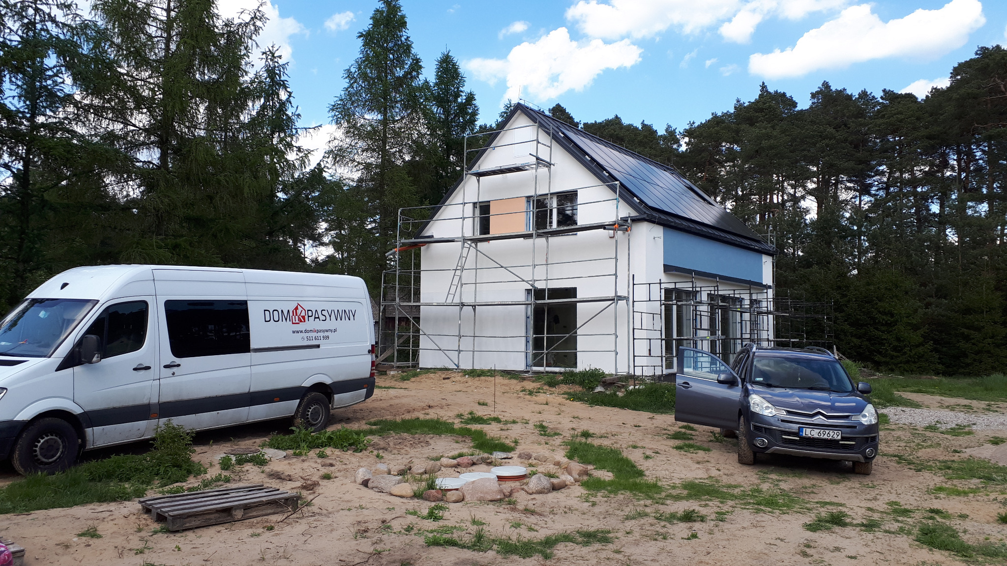 Dom w trakcie ocieplania, z rusztowaniem i samochodem firmy. Widoczny fragment elewacji w kolorze białym i niebieskim. W tle las.