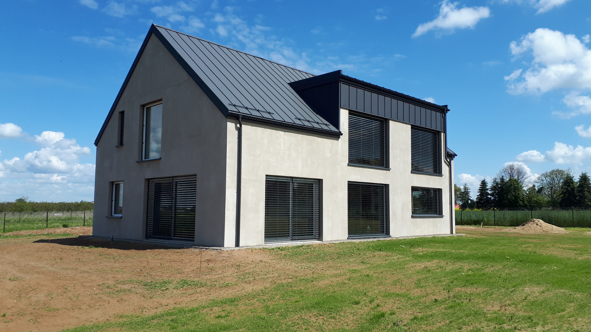Nowoczesny dom z szarą blachą na dachu, jasną elewacją i ciemnymi żaluzjami zewnętrznymi. Widok z zewnątrz, trawnik i błękitne niebo w tle.