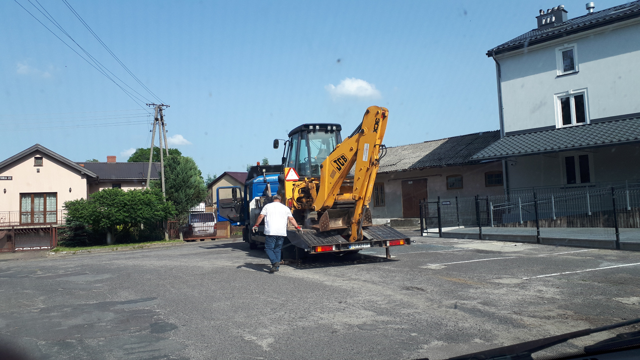 Żółta koparko-ładowarka JCB na niskopodwoziowej lawecie, przygotowywana do transportu. W tle budynki mieszkalne i słupy energetyczne.