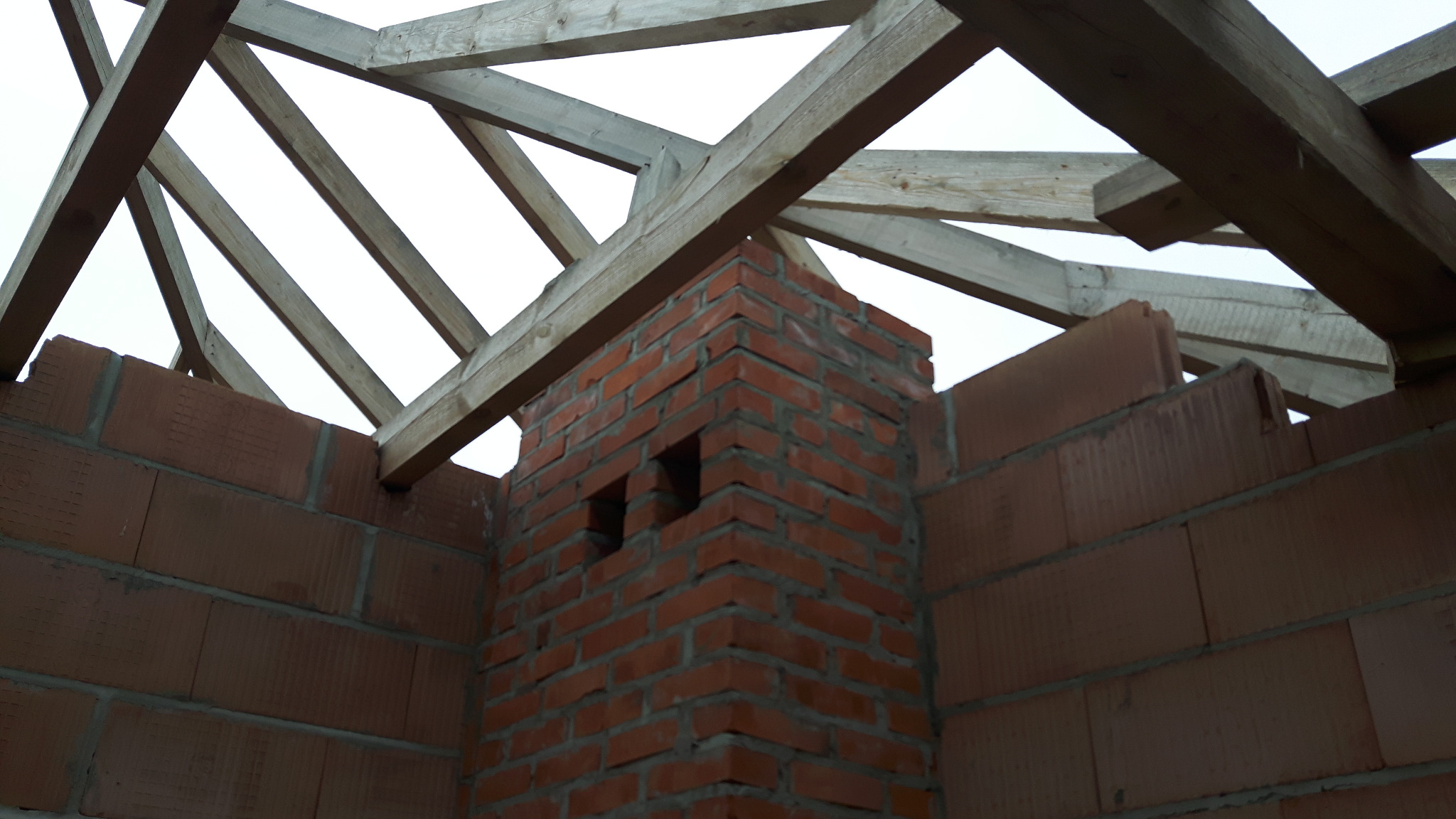 Wznoszenie komina z cegły klinkierowej na tle drewnianej konstrukcji dachu i ścian z pustaków ceramicznych. Widoczne otwory wentylacyjne w kominie.