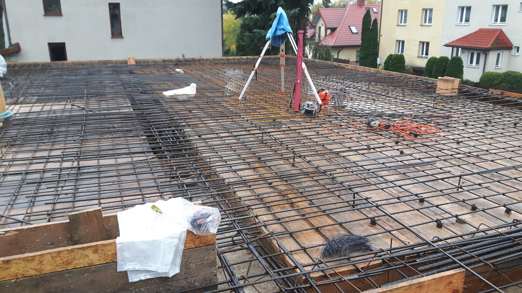 Zbrojenie stropu na deskowaniu z widocznym niwelatorem i narzędziami. W tle budynki mieszkalne.