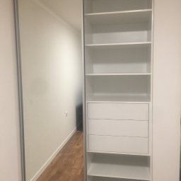 Andrii Pavlov - Biała szafa wnękowa z przesuwnymi drzwiami lustrzanymi, widoczne półki i szuflady, drewniana podłoga odbija się w lustrze, minimalistyczny design.