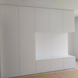 Andrii Pavlov - Biała zabudowa meblowa na wymiar z szafkami i otwartą przestrzenią roboczą, widok na jasne wnętrze z parkietem i grzejnikiem. Minimalistyczny design.