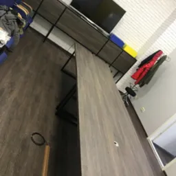 Nowoczesne biuro z długim stołem konferencyjnym o drewnianym wykończeniu, szafką RTV z telewizorem i białymi ceglanymi ścianami. Widok z perspektywy.