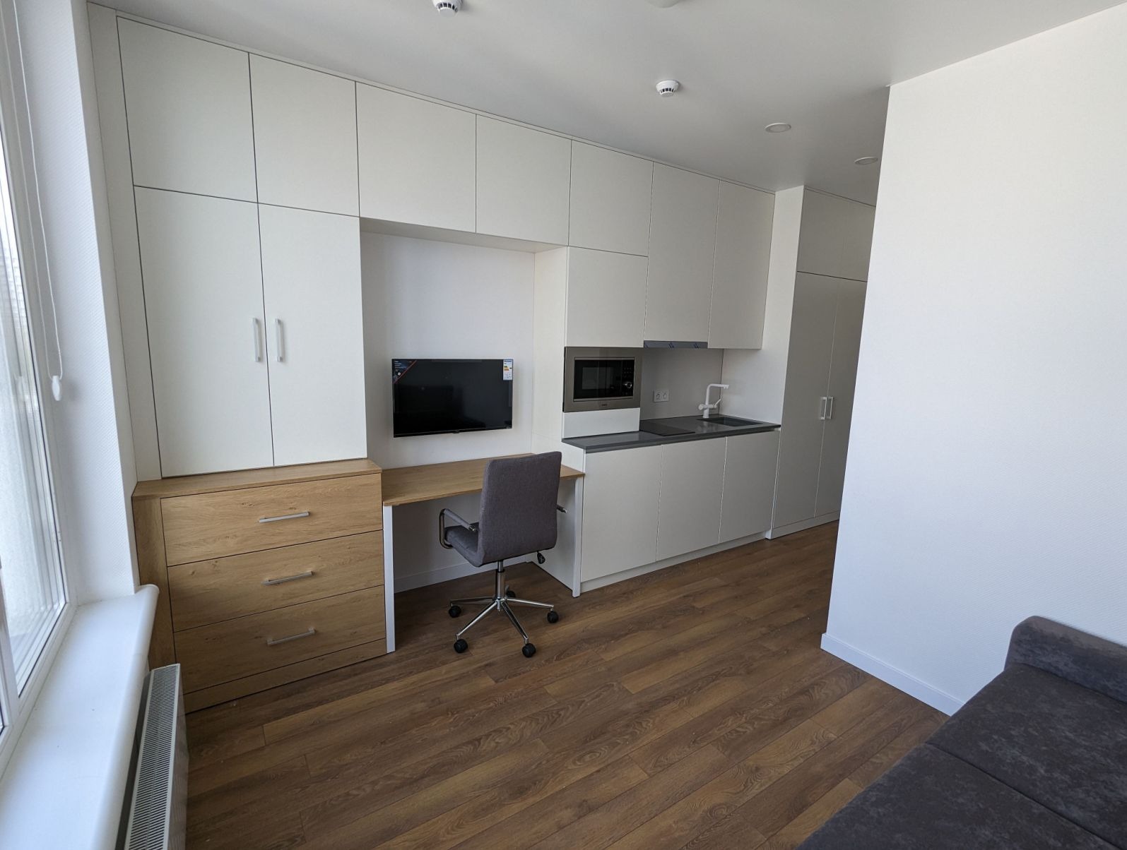 Nowoczesne studio z zabudową meblową: białe szafy, drewniana komoda z biurkiem, aneks kuchenny z szarym blatem i krzesło biurowe na tle drewnianej podłogi.