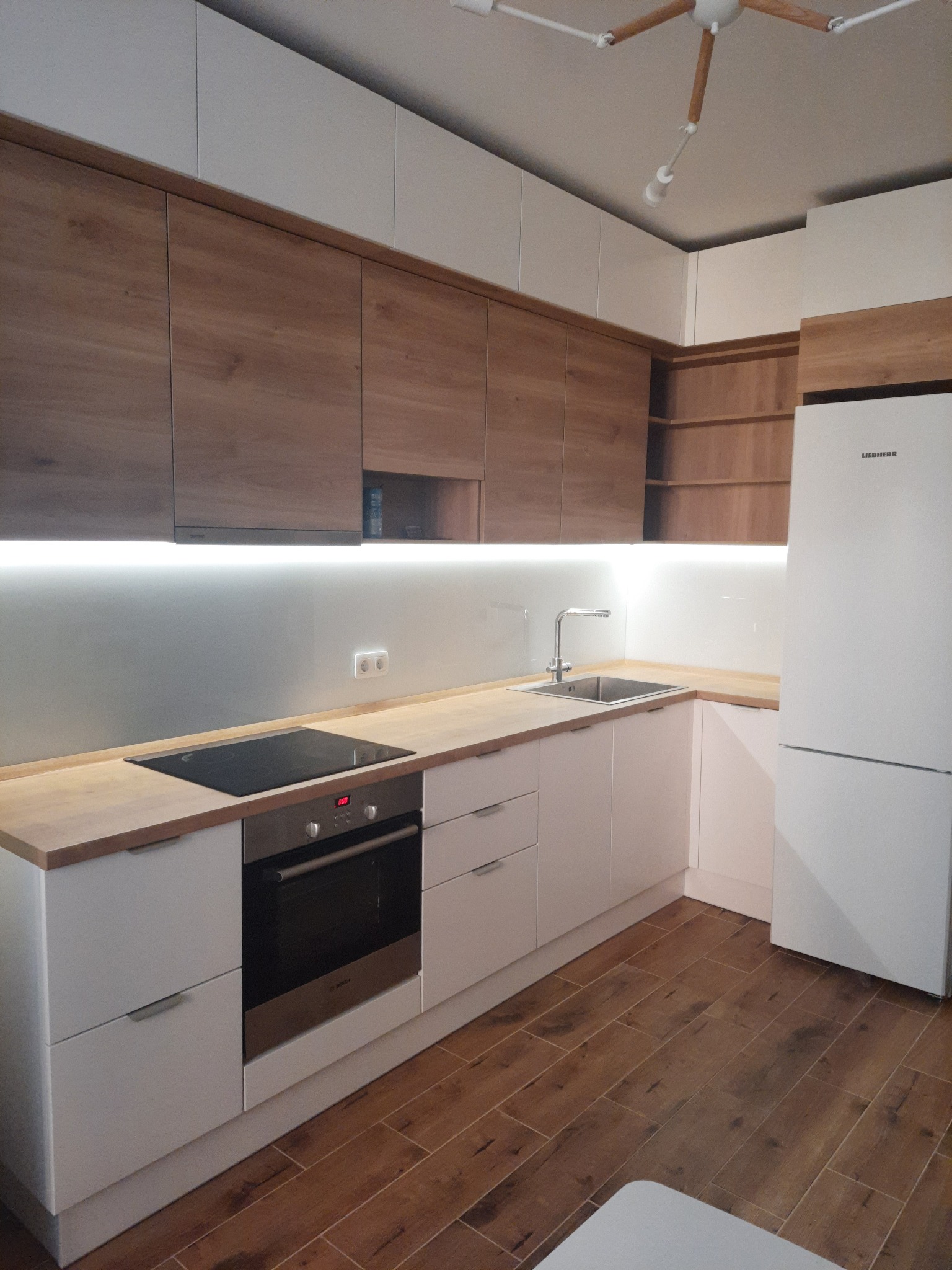 Nowoczesna kuchnia z drewnianymi szafkami górnymi i białymi dolnymi. Blat w kolorze drewna, oświetlenie LED podszafkowe, brązowa podłoga imitująca drewno.