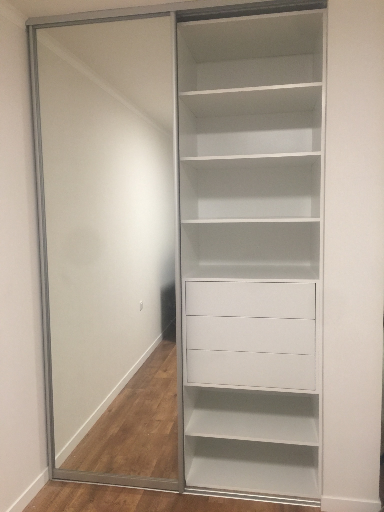 Biała szafa wnękowa z przesuwnymi drzwiami lustrzanymi, widoczne półki i szuflady, drewniana podłoga odbija się w lustrze, minimalistyczny design.