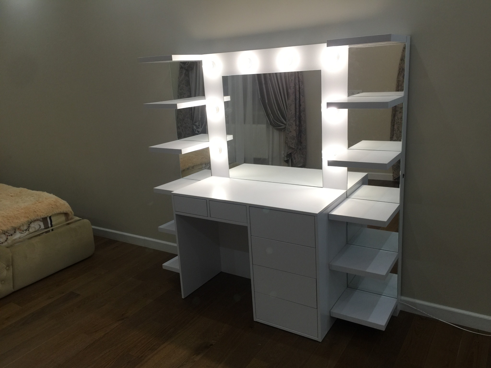 Biała toaletka z lustrem i oświetleniem LED, z półkami po bokach, na tle ściany i fragmentu łóżka. Widoczne detale wykończenia i układ półek.