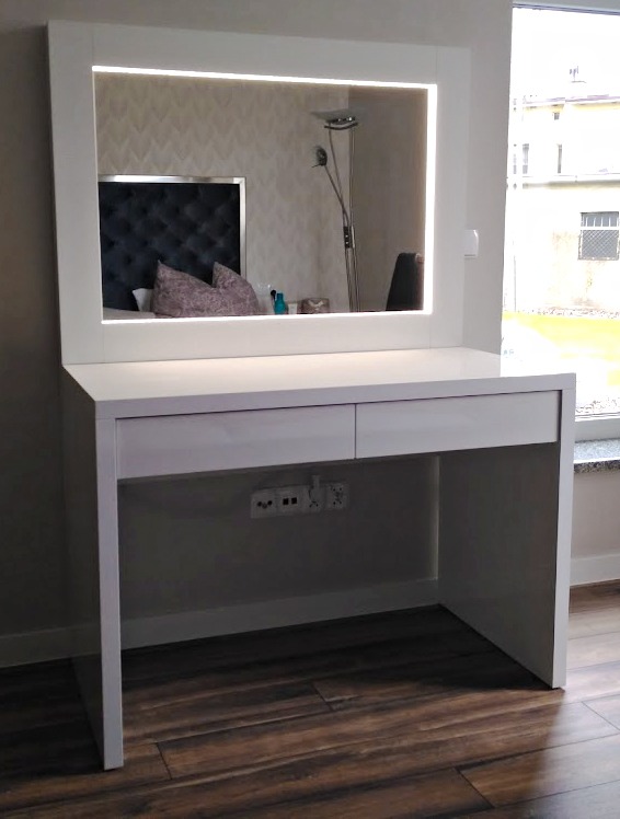 Biała toaletka z podświetlanym lustrem w nowoczesnym stylu, odbijająca fragment sypialni z pikowanym zagłówkiem i lampą podłogową. Ujęcie na tle okna.