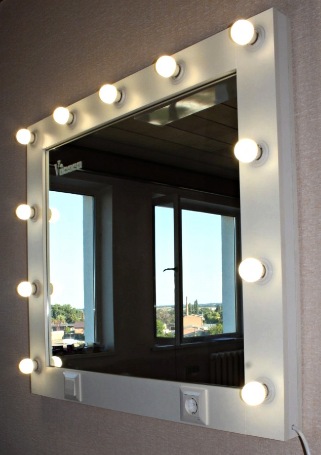 Białe lustro do makijażu z żarówkami na ramie i gniazdkami, odbijające okno z widokiem na zieleń, zamontowane na ścianie.