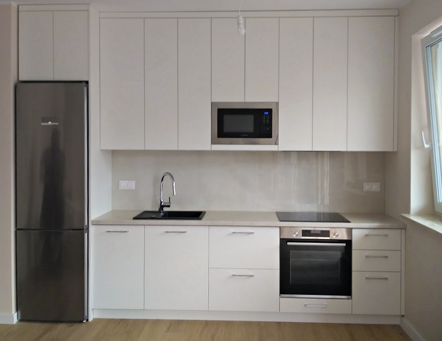 Biała, nowoczesna kuchnia z zabudową na wymiar: szafki wiszące i stojące, piekarnik, płyta indukcyjna, zlew, bateria i lodówka. Minimalistyczny design, jasne wnętrze.