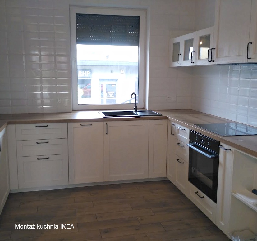 Jasna kuchnia z białymi frontami, czarnymi uchwytami i drewnianym blatem. Widoczne okno z roletą i czarny zlew. Na podłodze panele imitujące drewno.