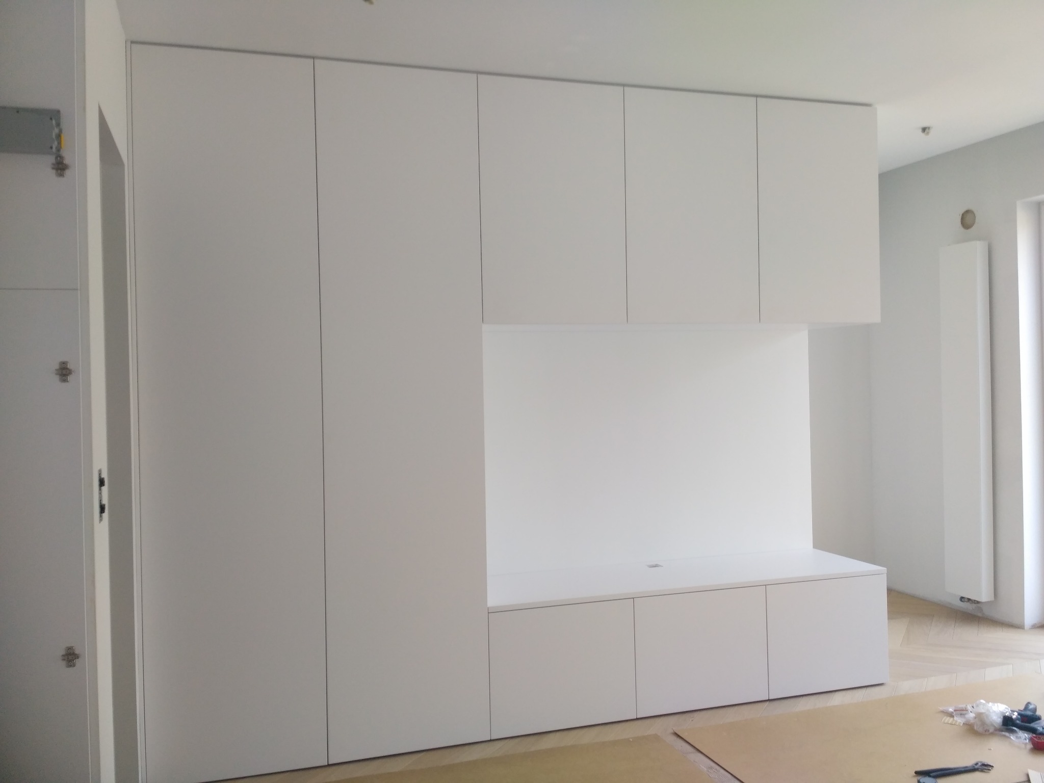 Biała zabudowa meblowa na wymiar z szafkami i otwartą przestrzenią roboczą, widok na jasne wnętrze z parkietem i grzejnikiem. Minimalistyczny design.