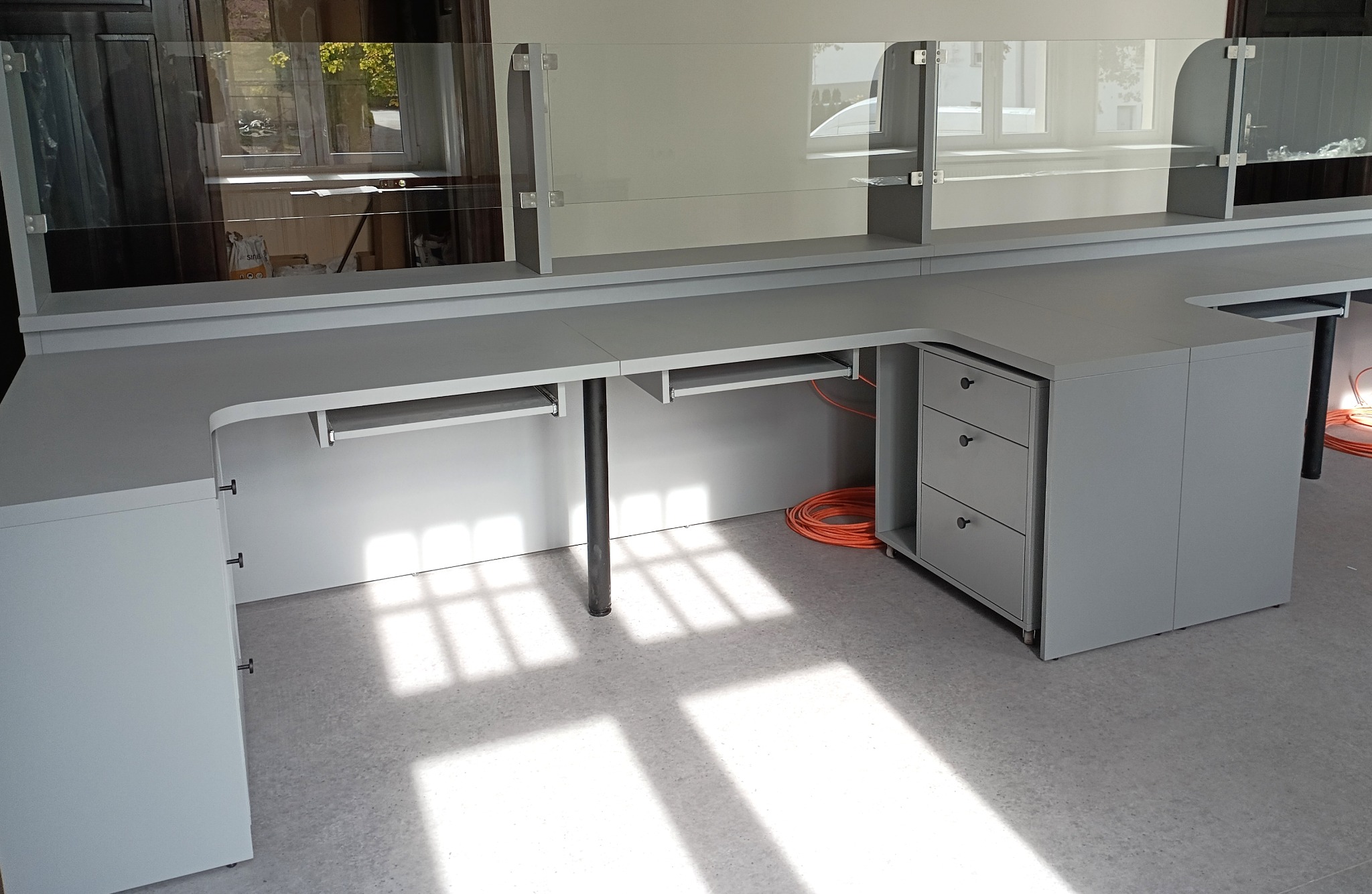 Szara, narożna szafa biurowa na wymiar z szklaną nadstawką, widok z boku,  z elementami szuflad i wysuwanymi półkami na klawiaturę, kabel w tle.