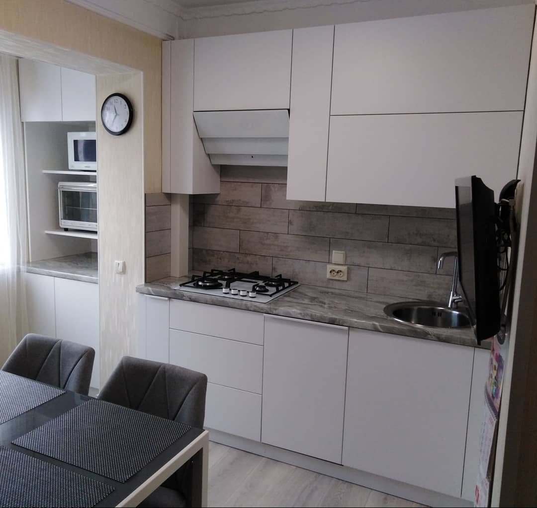 Nowoczesna, jasna kuchnia z białymi szafkami, szarym blatem i płytkami imitującymi drewno na ścianie. Widoczny fragment stołu z krzesłami i kuchenka gazowa.