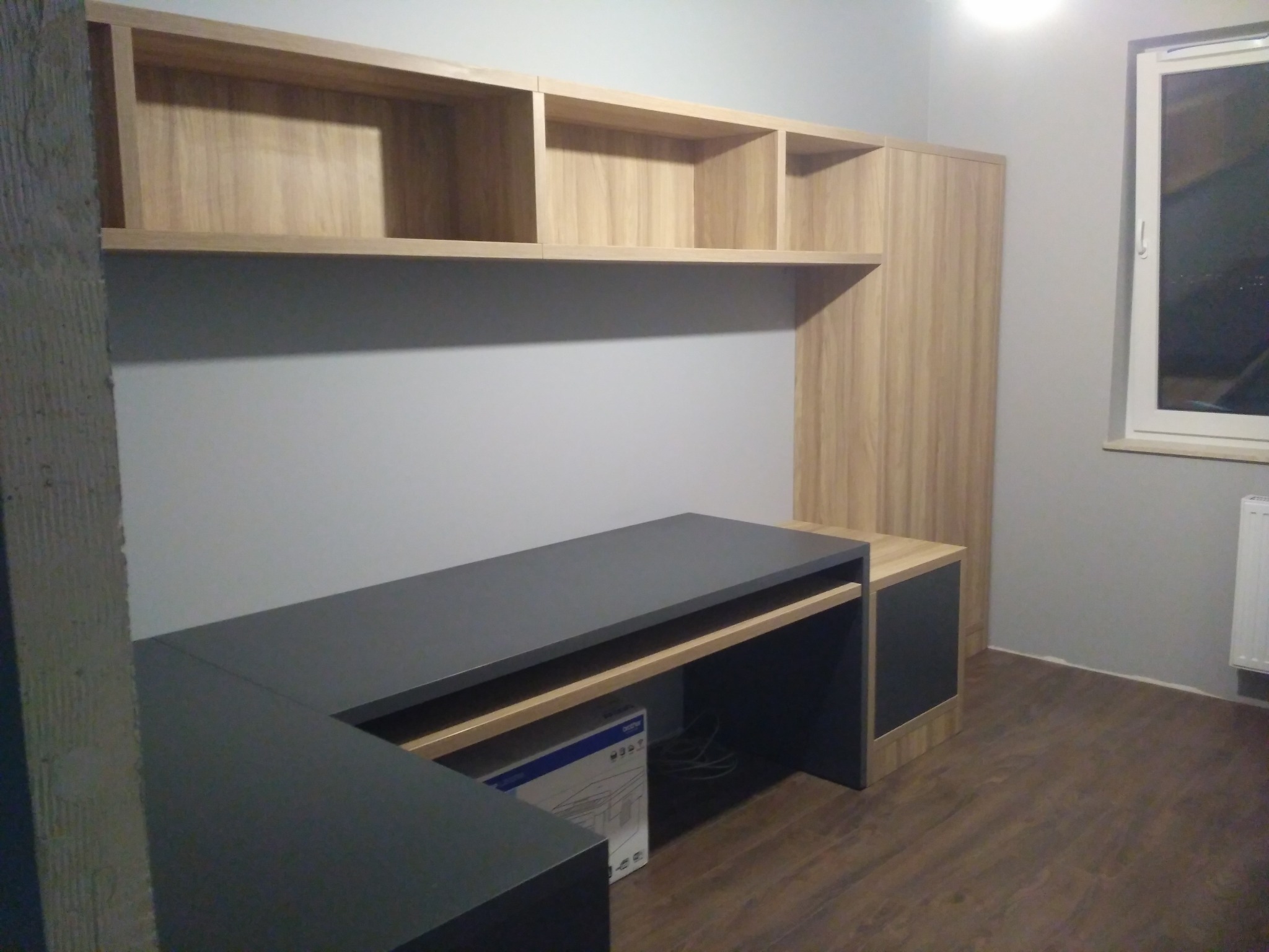 Nowoczesna szafa wnękowa z biurkiem i półkami w odcieniach drewna i szarości, z widocznym fragmentem ściany i podłogi, tworząca spójną całość.