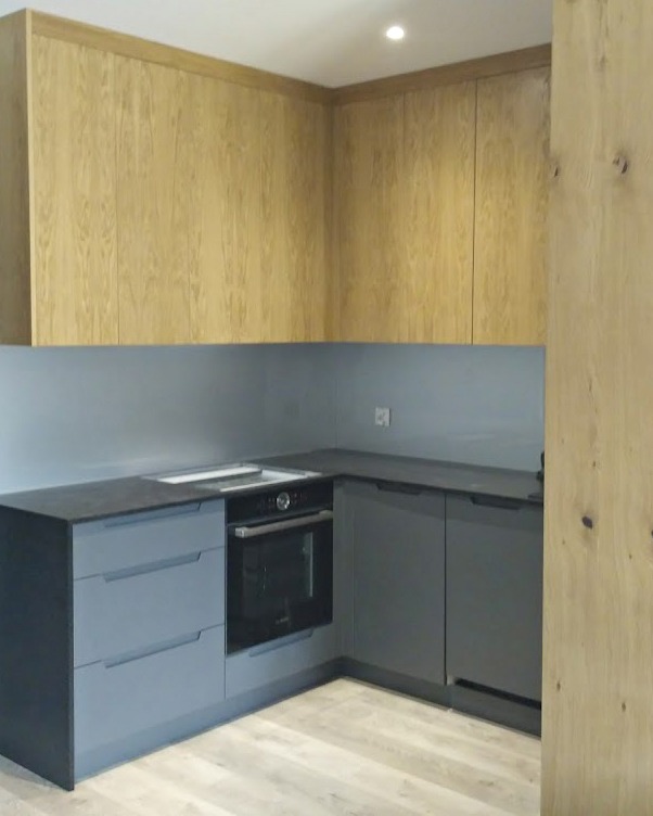 Nowoczesna kuchnia narożna z szarymi szafkami dolnymi i jasnymi, drewnianymi szafkami górnymi, z piekarnikiem w zabudowie i jasną podłogą.