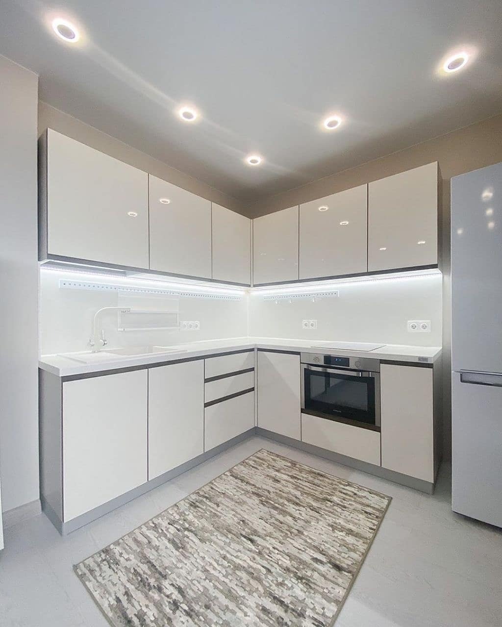 Nowoczesna kuchnia w bieli z szarymi akcentami i dywanem. Górne szafki podświetlone LED, minimalistyczny design, oświetlenie punktowe sufitowe i sprzęt AGD.