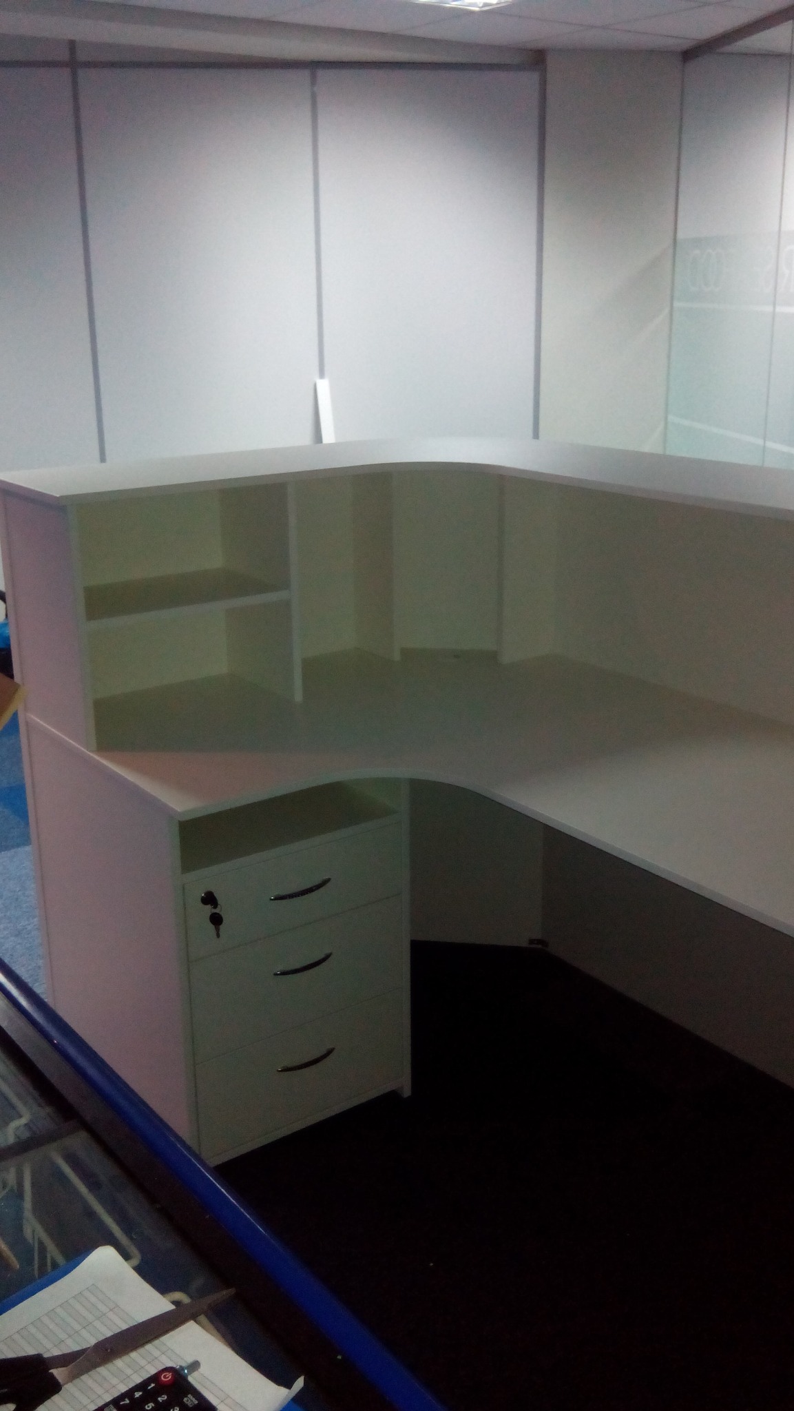 Biała, narożna lada recepcyjna z szufladami i półkami, widoczna w przestrzeni biurowej z panelami ściennymi. Klawiatura i dokumenty na pierwszym planie.