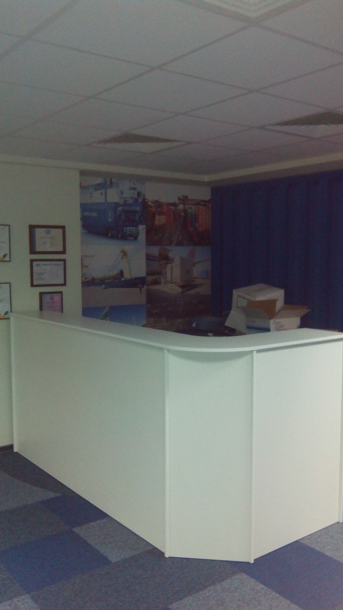 Biała lada recepcyjna z zaokrąglonym blatem w biurze, na tle ściany z grafiką transportową i dywanem w geometryczne wzory. Widoczne certyfikaty na ścianie.