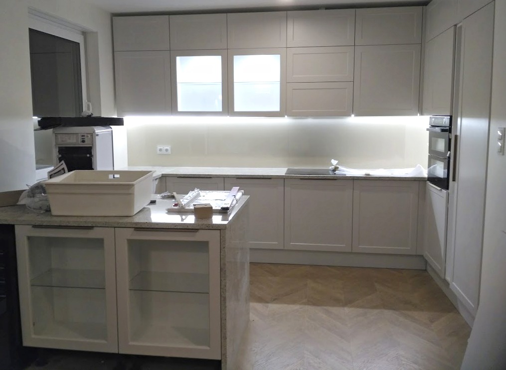 Jasna kuchnia z białymi szafkami i podświetleniem LED pod szafkami wiszącymi. Na wyspie kuchennej leżą narzędzia montażowe. Parkiet ułożony w jodełkę.