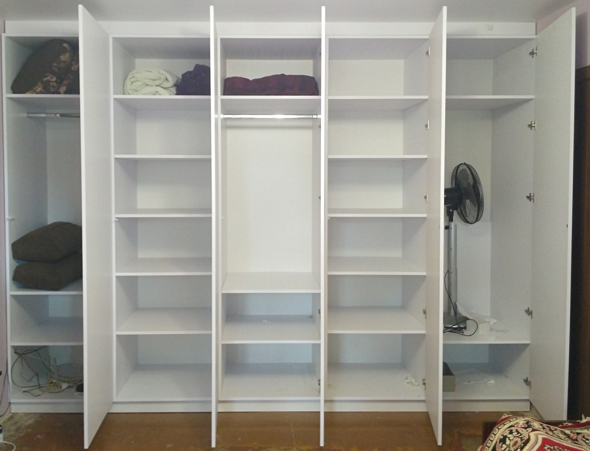 Biała szafa wnękowa z otwartymi drzwiami, widoczne półki i ubrania. W jednej z komór stoi wentylator. Podłoga brązowa, w tle fragment wzorzystej tkaniny.