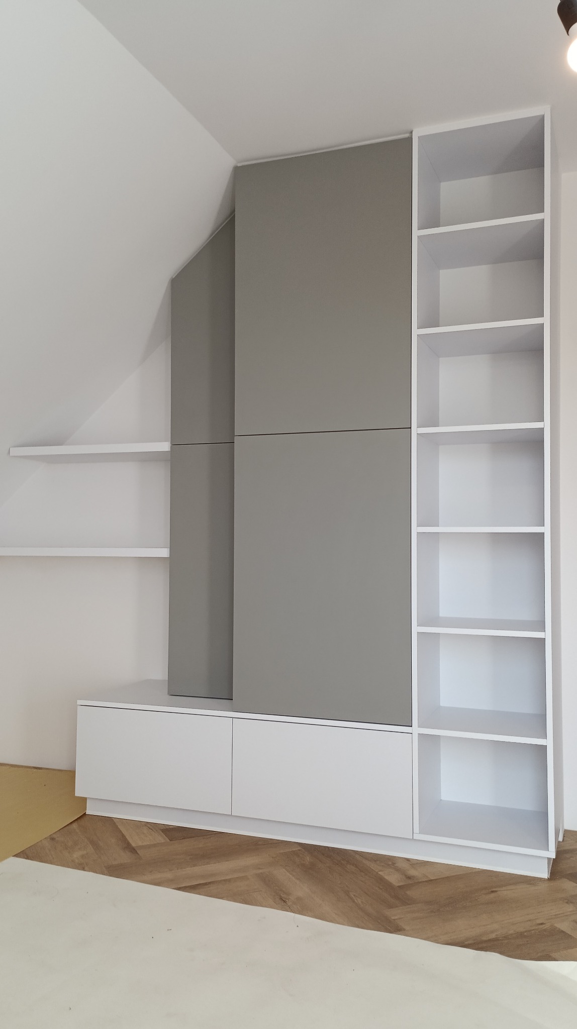 Szafa wnękowa z szarymi frontami i białymi półkami, dopasowana do skosu dachu. Widoczne białe półki wiszące i podłoga w jodełkę. Minimalistyczny design.