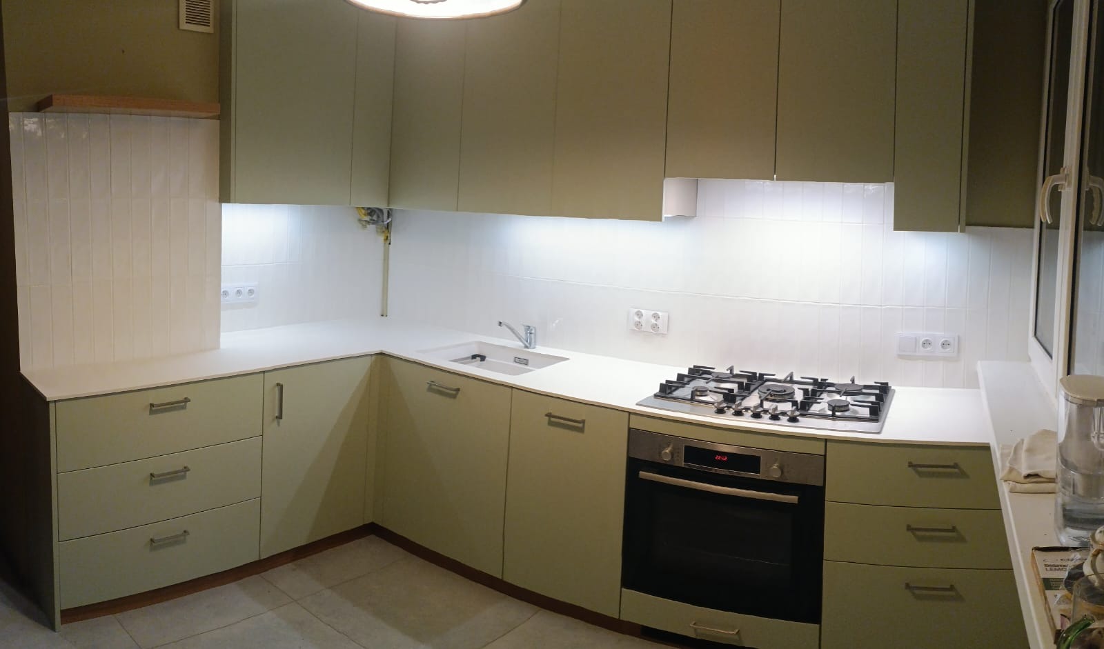 Jasnozielona kuchnia z białym blatem i płytkami. Widoczna płyta gazowa, piekarnik i zlew. Górne szafki podświetlone. Minimalistyczny design, nowoczesne wykończenie.