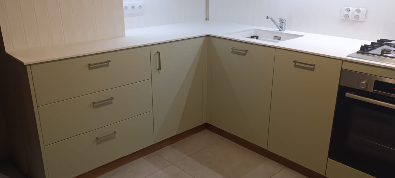 Jasna kuchnia na wymiar z beżowymi frontami, srebrnymi uchwytami i białym blatem. Widoczny zlew, płyta grzewcza i piekarnik. Ściana wykończona białą boazerią.