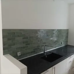 Kuchnia z zieloną, ceglaną mozaiką nad czarnym blatem i białymi szafkami, widoczny kran i okno.